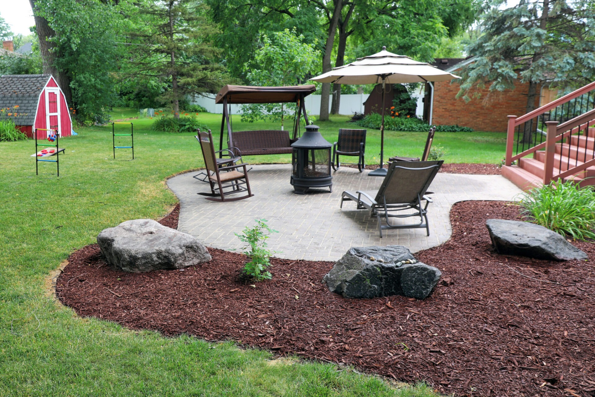 A serene backyard scene with patio chairs, a fire pit, and a swing beneath an umbrella. Red shed and lawn games in the background.