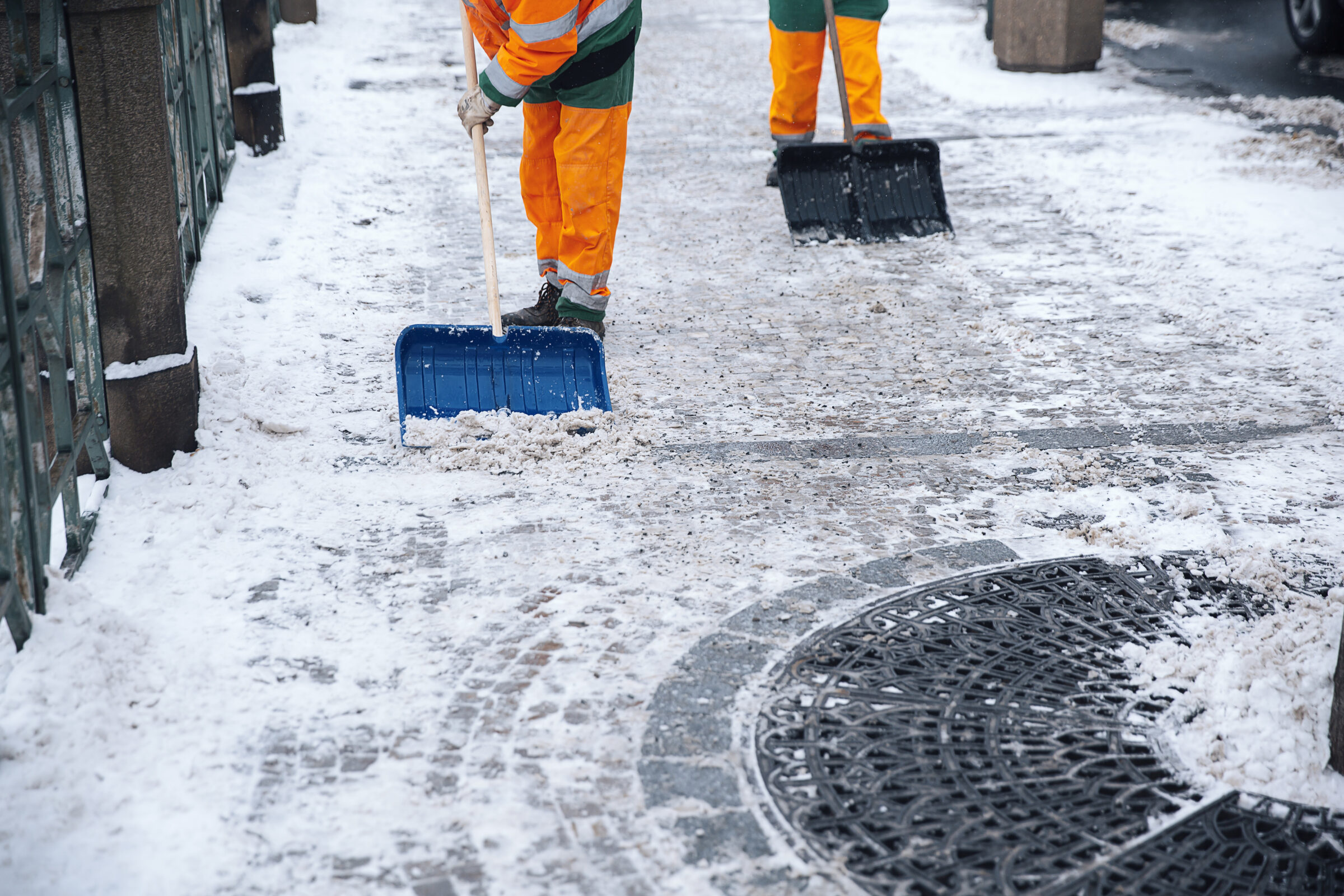 Two people in orange gear shovel snow on a brick sidewalk, clearing a path in a cold, urban environment.