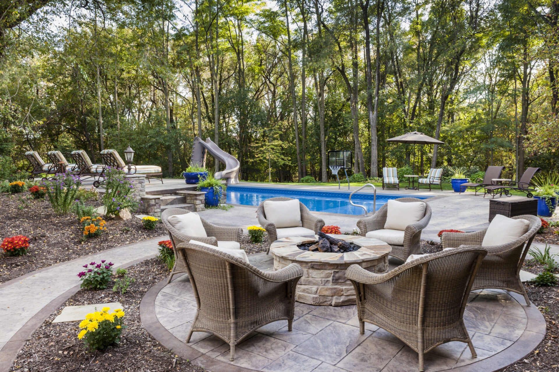 Backyard oasis featuring a circular fire pit, wicker chairs, garden, pool with slide, and loungers surrounded by lush trees and vibrant flowers.