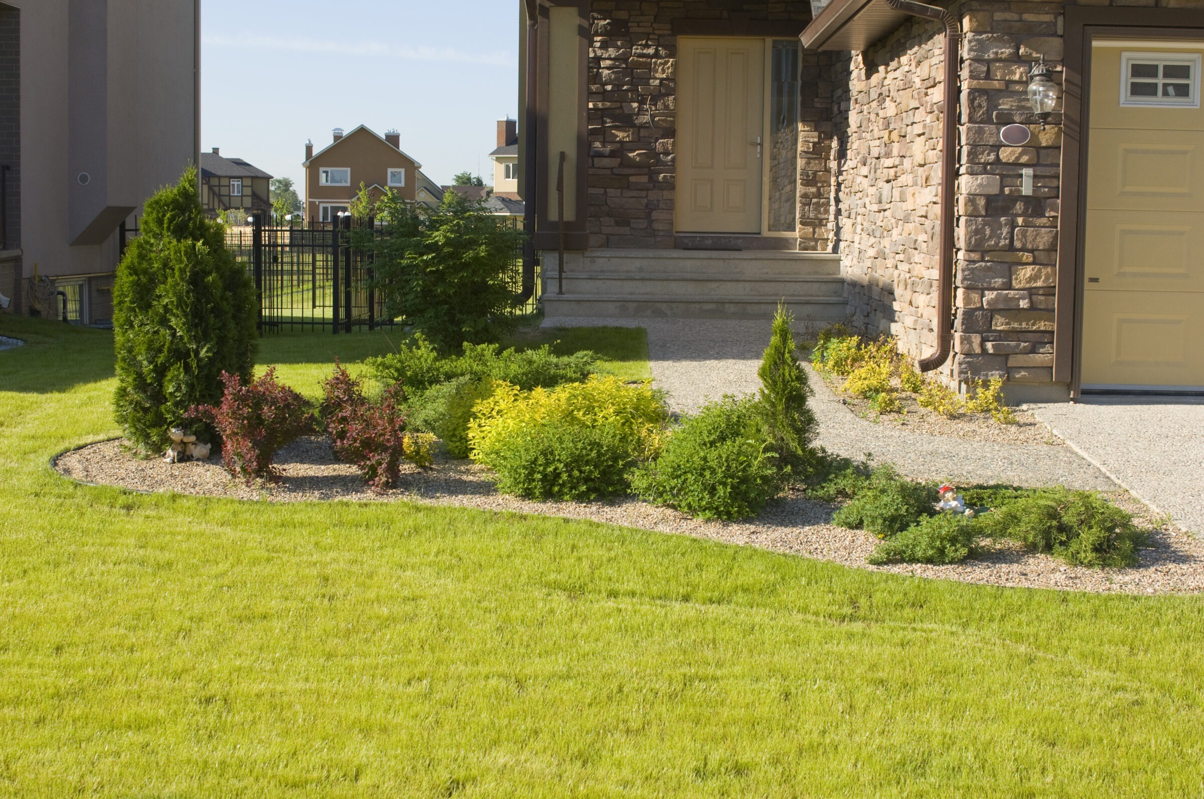 A well-maintained garden in front of a stone house, featuring green shrubs, a paved pathway, and neatly trimmed grass in a suburban setting.
