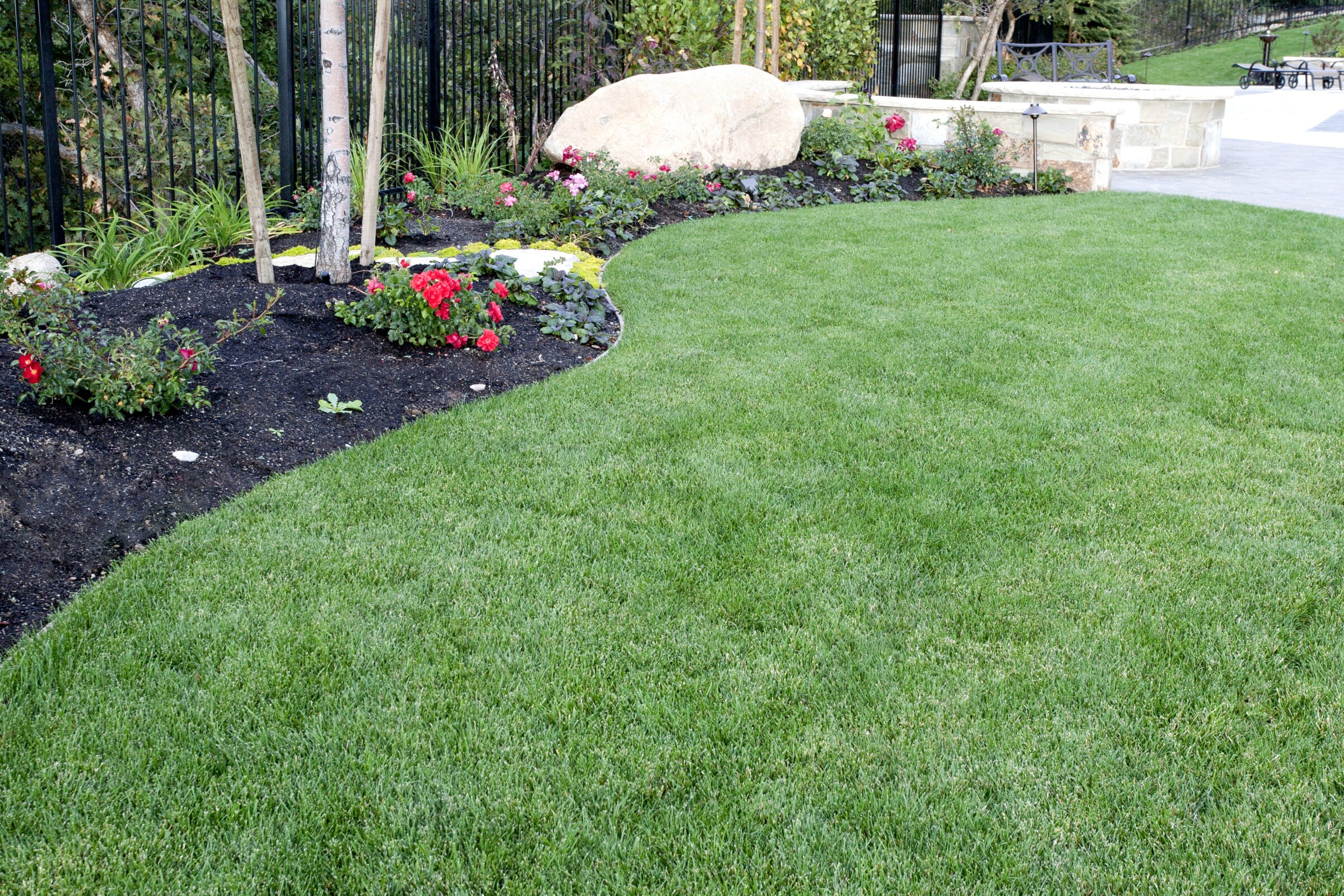 A landscaped garden features lush green grass, colorful flowers, small trees, and a large rock. Fenced area with patio chairs is visible.