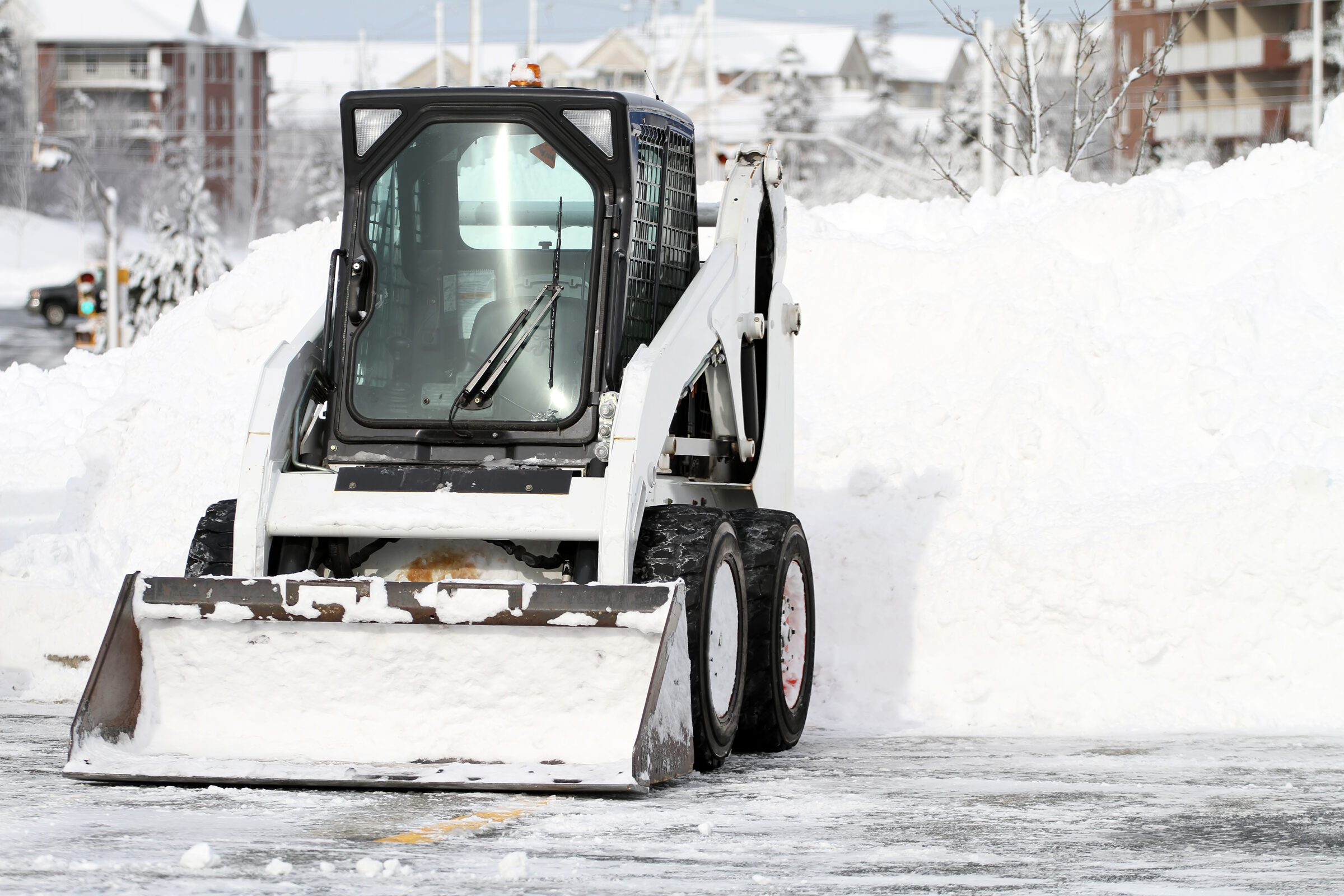 A skid-steer loader removes snow near residential buildings in a snowy urban area, with trees and a cloudy sky in the background.