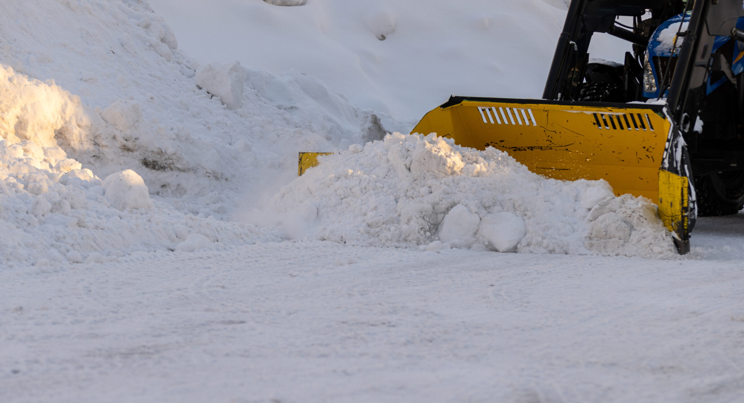 A snowplow clears a path through deep, icy snow on a winter day, surrounded by large snowdrifts and a clear, cold atmosphere.