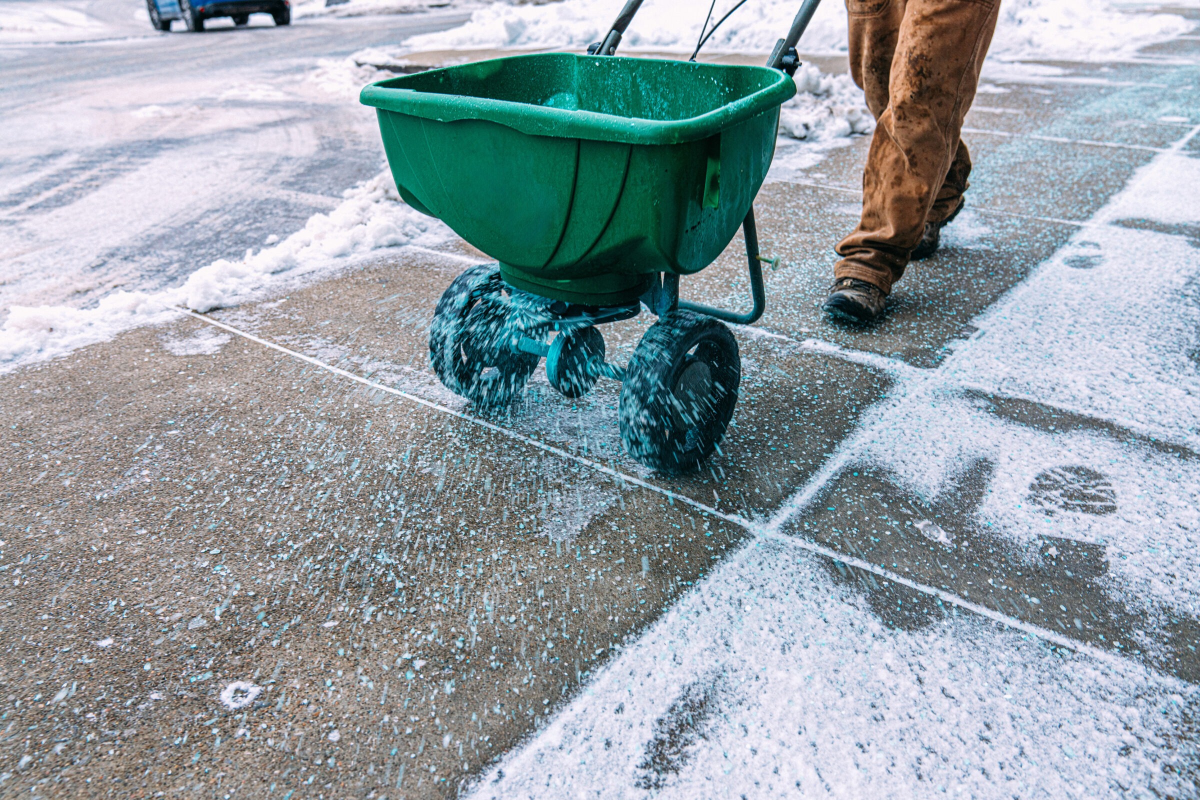 A person uses a green spreader to distribute salt on a snowy sidewalk, preventing ice. Nearby road is lightly covered in snow.