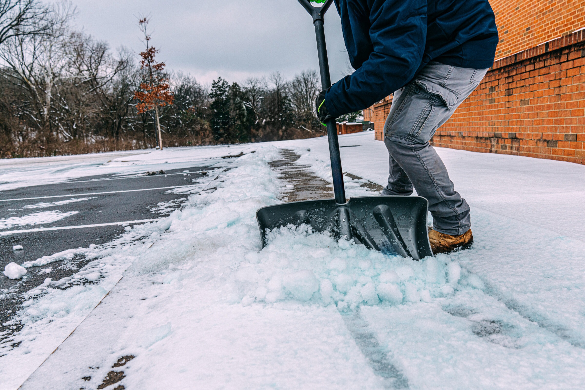 A person in winter clothing shovels snow from a pathway next to a brick building, surrounded by trees and a cloudy sky.