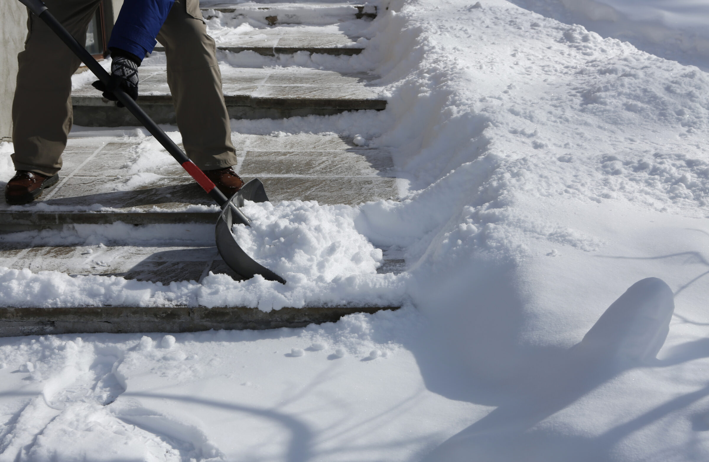 A person is shoveling snow from concrete steps on a sunny winter day, creating clear pathways in a snow-covered area.