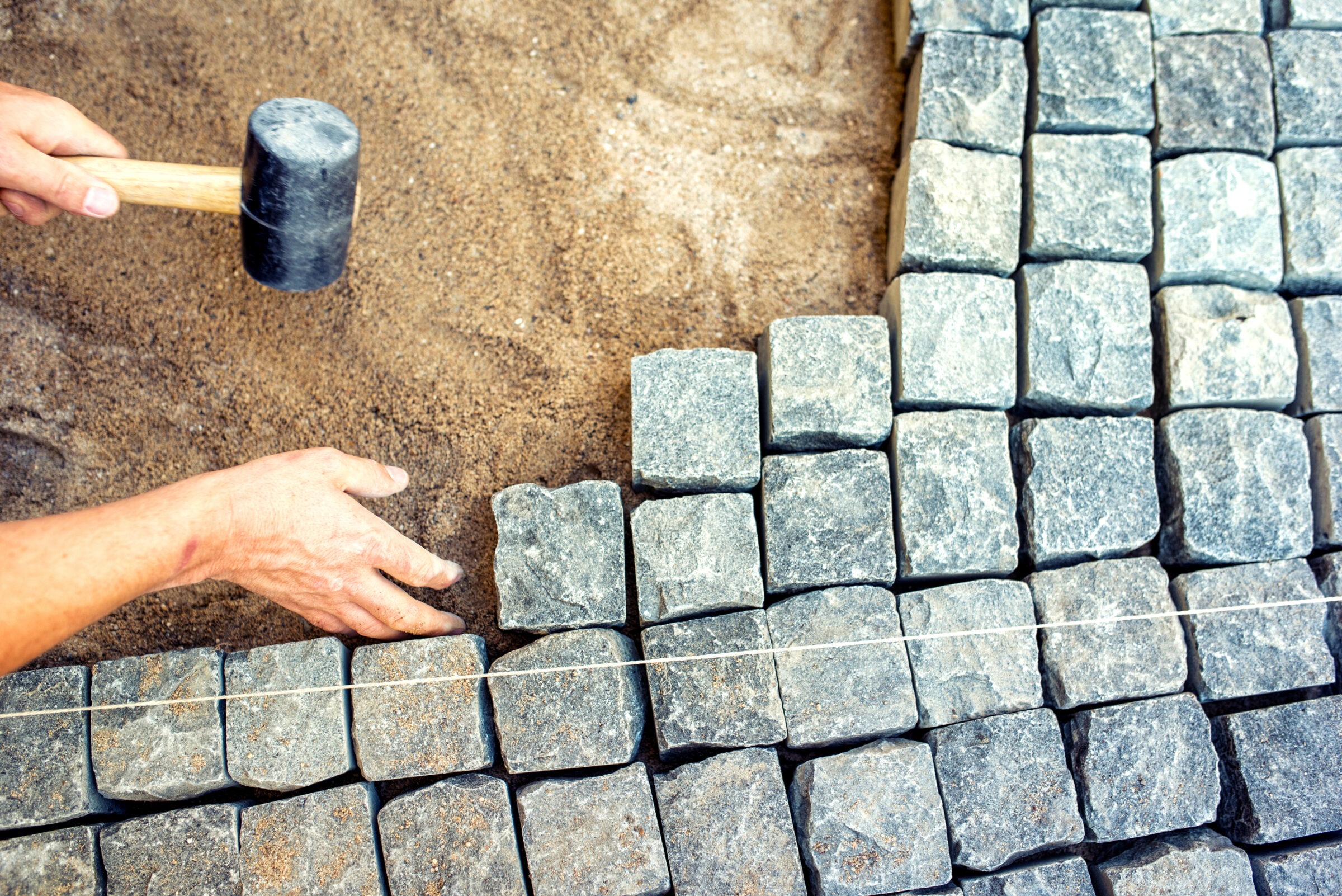 A person aligns stone pavers with a mallet on a sandy surface, using a string line for precision in arrangement.