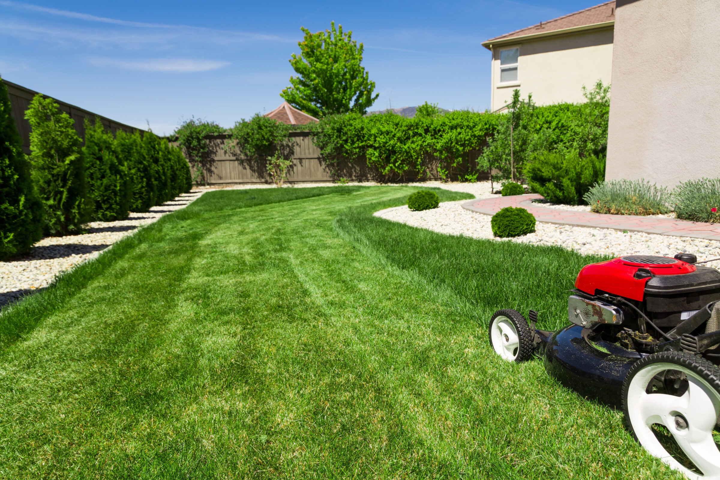 A red lawn mower sits on freshly cut grass, near a white house and wooden fence, under a clear blue sky.