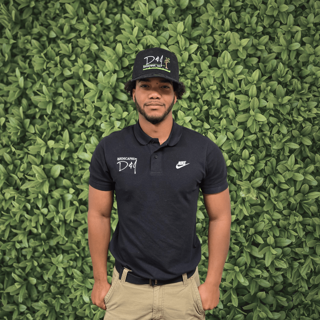 A person wearing a black polo and hat stands in front of a lush, green hedge, displaying logos on their attire.