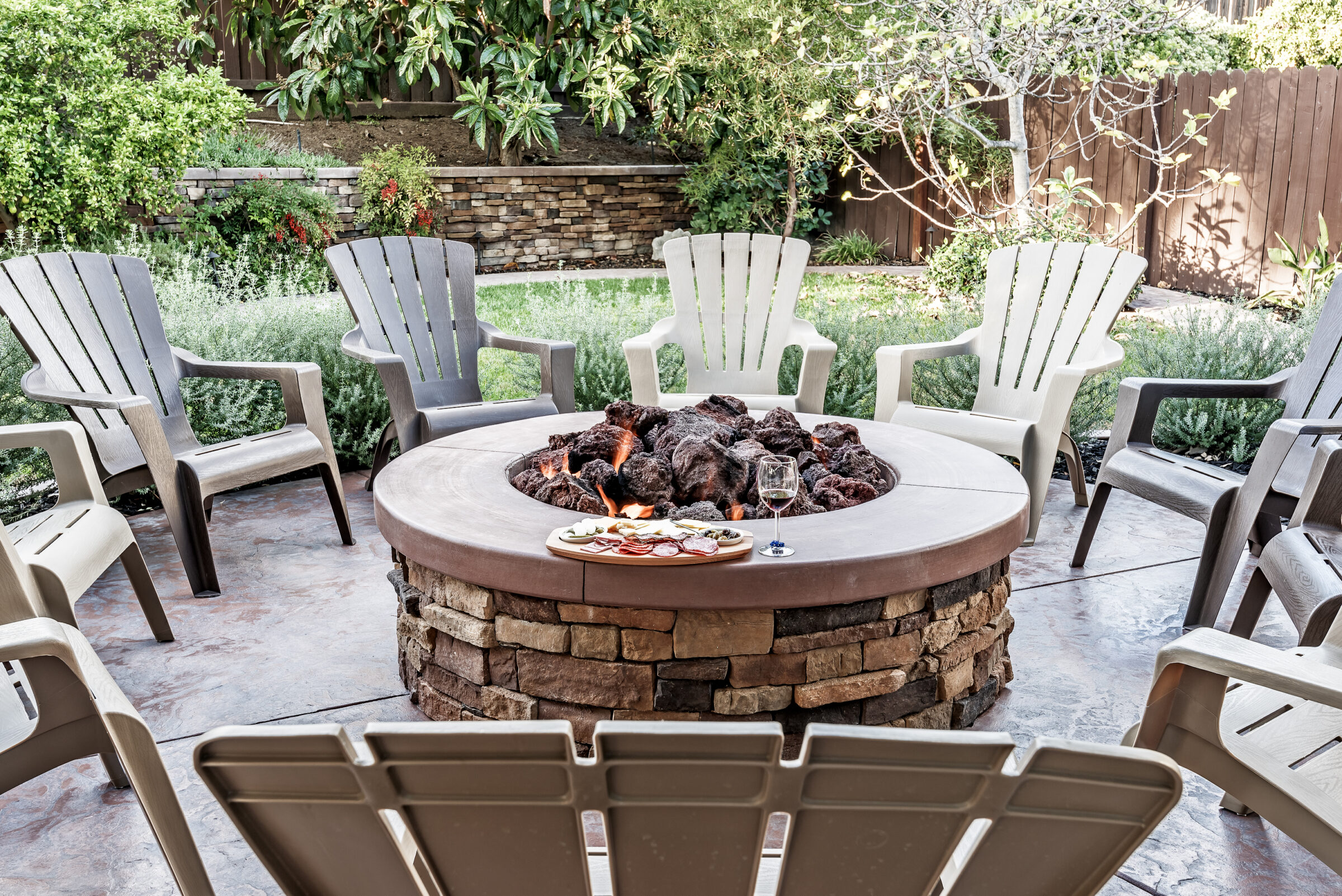 Large outdoor fire pit surrounded by wooden rocking chairs, beautifully landscaped backyard