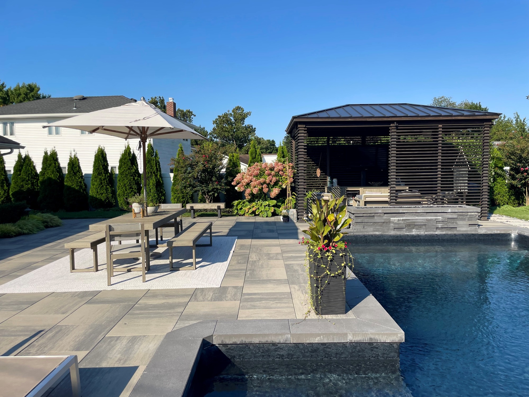 High-end, NY outdoor living space, a backyard patio with pergola, pavilion, cabana, chaise lounge chair, outdoor rug, tequila bar, dining set with table and chairs, couch, television, pool with waterfall, umbrella, fire pit and stunning grey patio. Modern vibe. Surrounded by lush cottage style gardens, planters, annuals, perennial flowers, shrubs and trees. Stunning entertainment space to be used all year round and to enjoy in the summer time. party, relax and unwind. Cool off in the pool, stay warm by the fire.