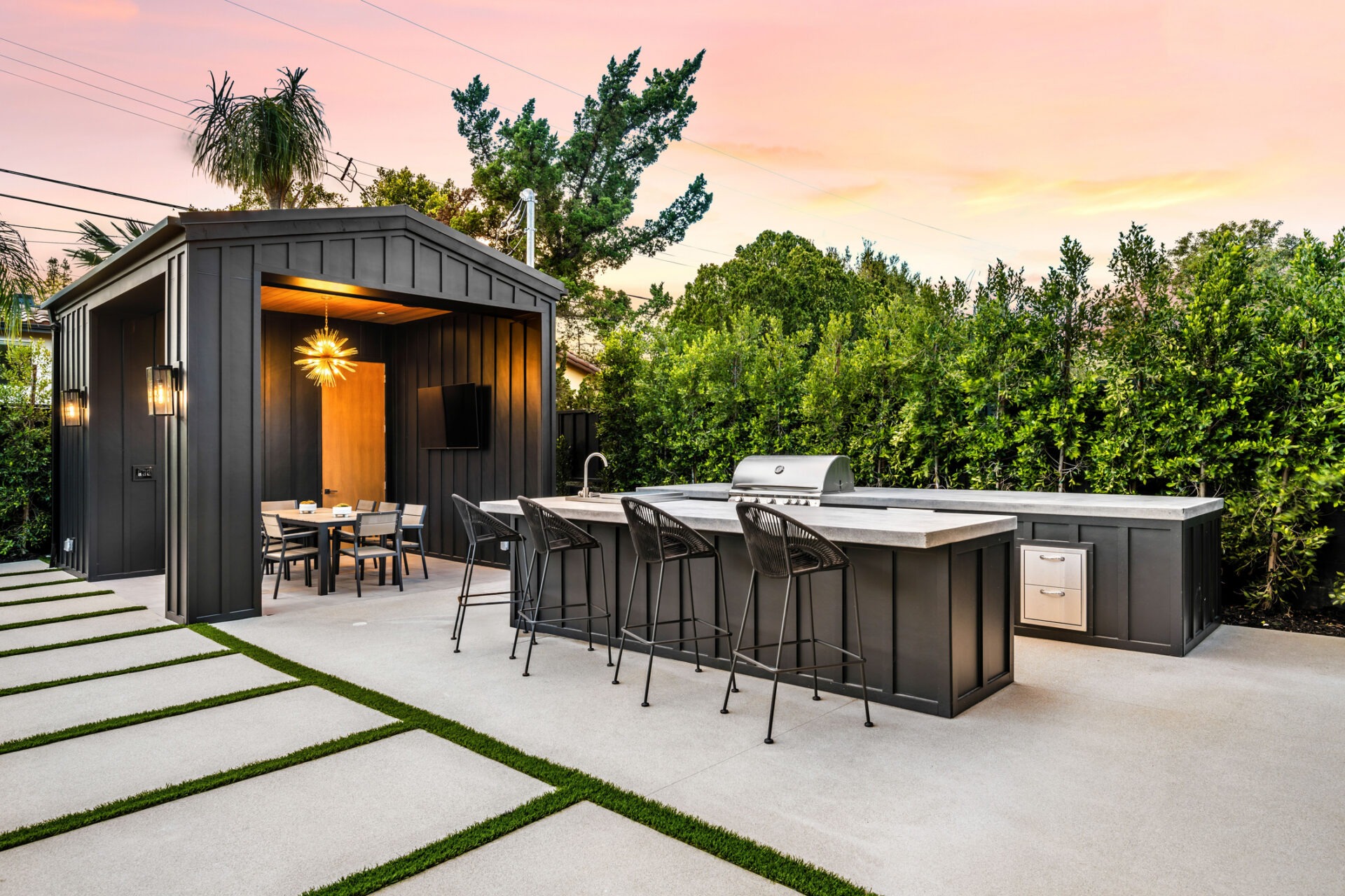 Modern outdoor kitchen and dining area with sleek design, featuring a grill, bar seating, and stylish lighting at sunset.